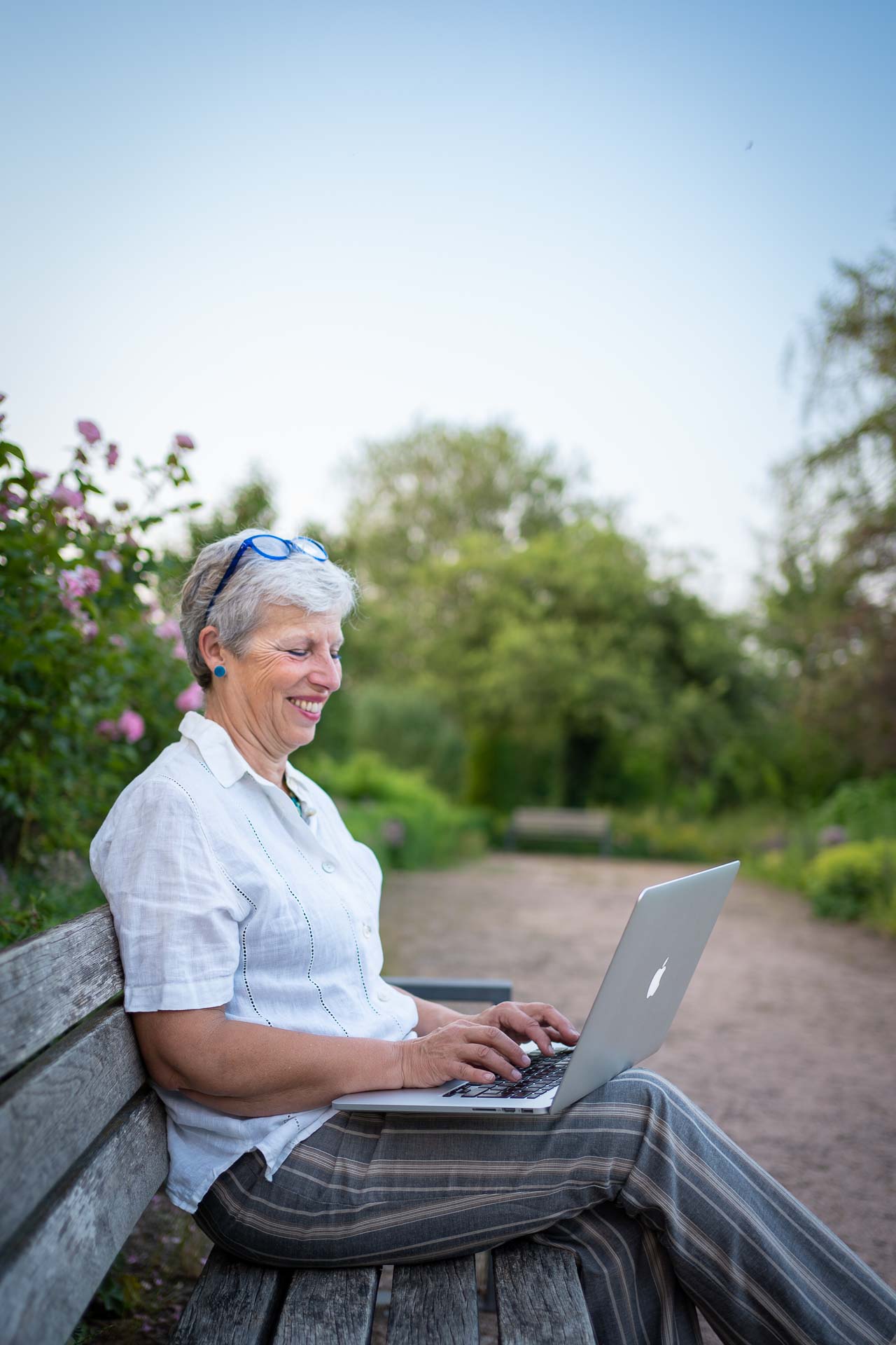 Christine Hembach sitzt auf einer Parkbank mit ihrem Laptop auf dem Schoß und schaut lachend auf ihr MacBook.